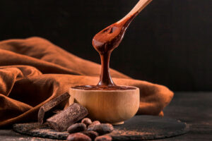 Vasilha com chocolate derretido com chocolate em barra e sementes de cacau ao lado representando visita à fábrica Nugali.
